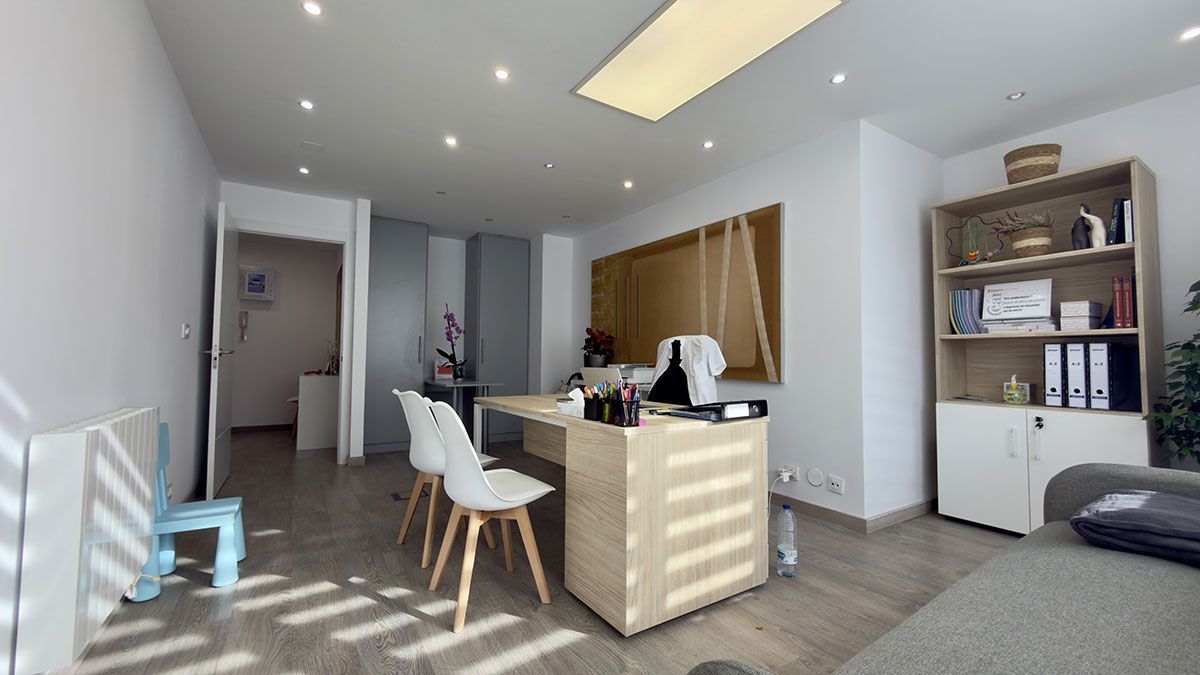 Una oficina luminosa con un escritorio, dos sillas blancas, una estantería de madera y una puerta que da a una habitación separada.