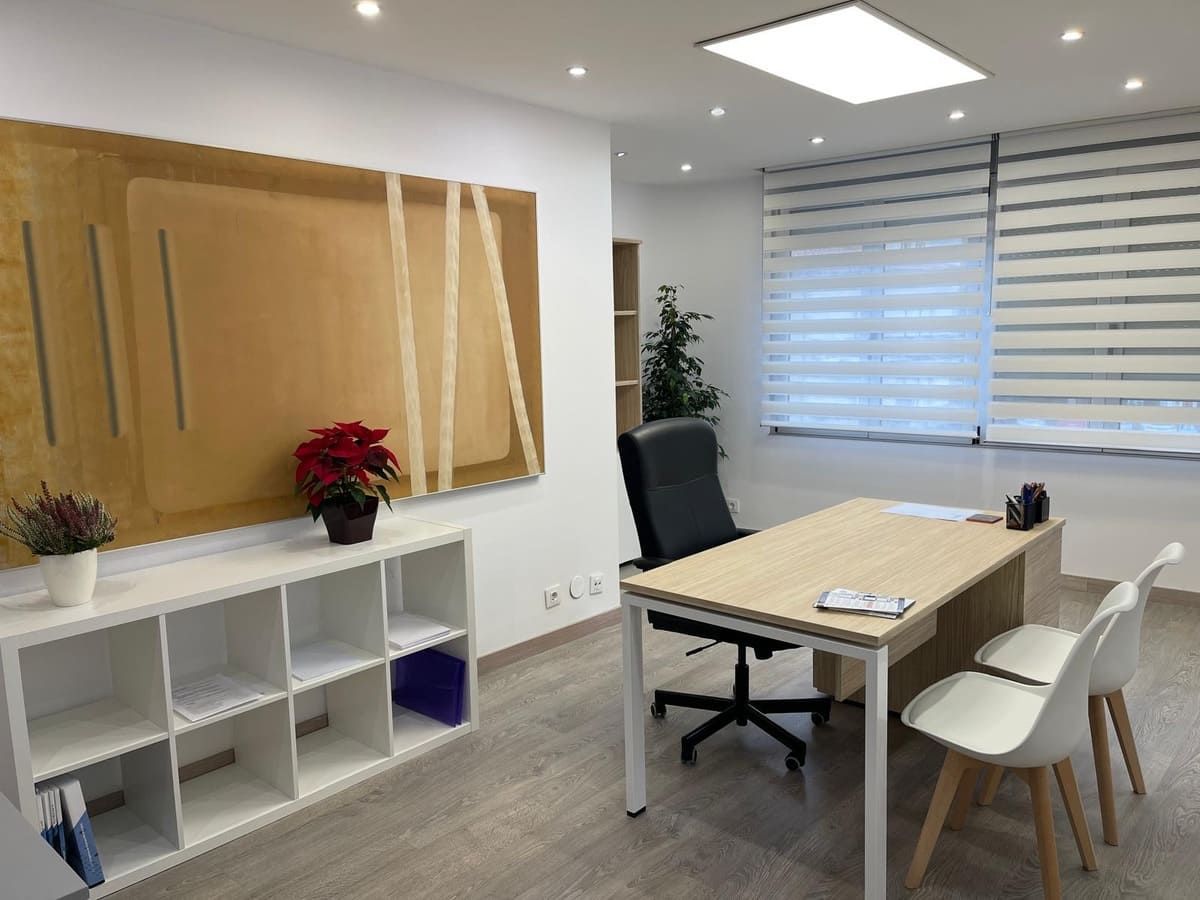 Interior de oficina con escritorio, sillas, obras de arte y una ventana con persianas.