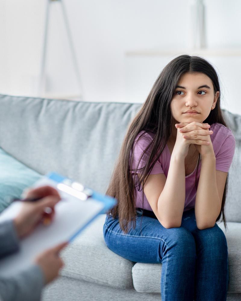 Adolescente sentada en un sofá, con expresión pensativa y las manos entrelazadas. En primer plano, una persona sostiene una carpeta.