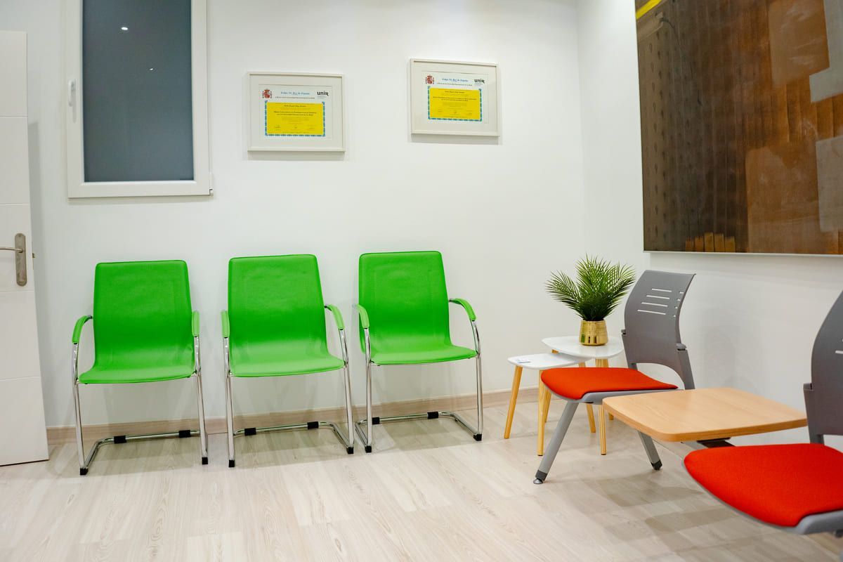 Sala de espera con sillas verdes y rojas contra paredes blancas; certificados enmarcados, obras de arte y una mesita con flores.