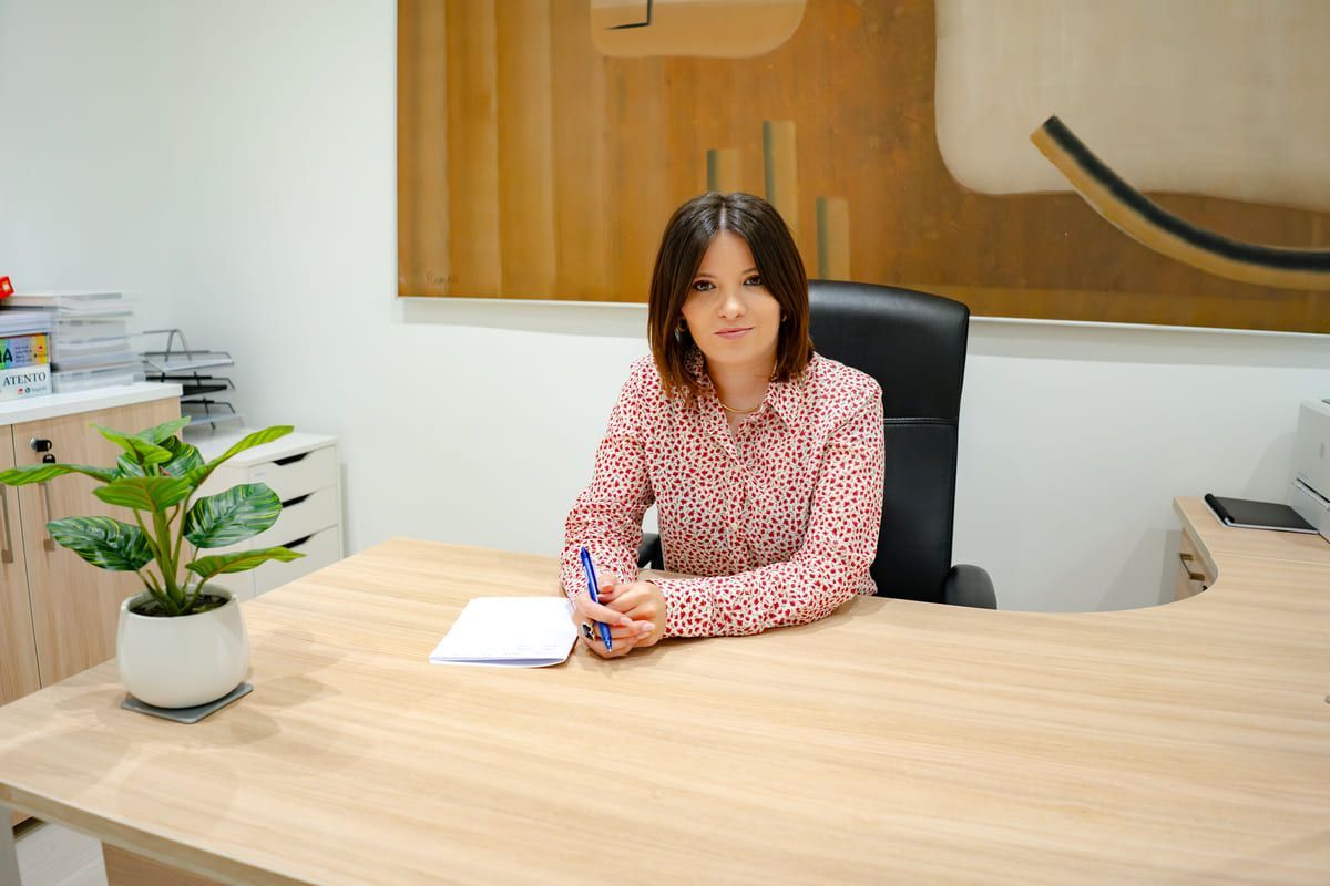Una mujer con una blusa floreada está sentada en un escritorio en una oficina, mirando a la cámara.