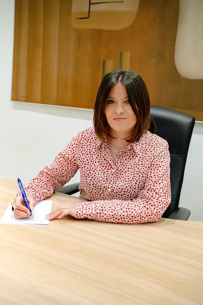Mujer con camisa roja de lunares, sentada a un escritorio, escribiendo con un bolígrafo. Obra de arte en la pared.