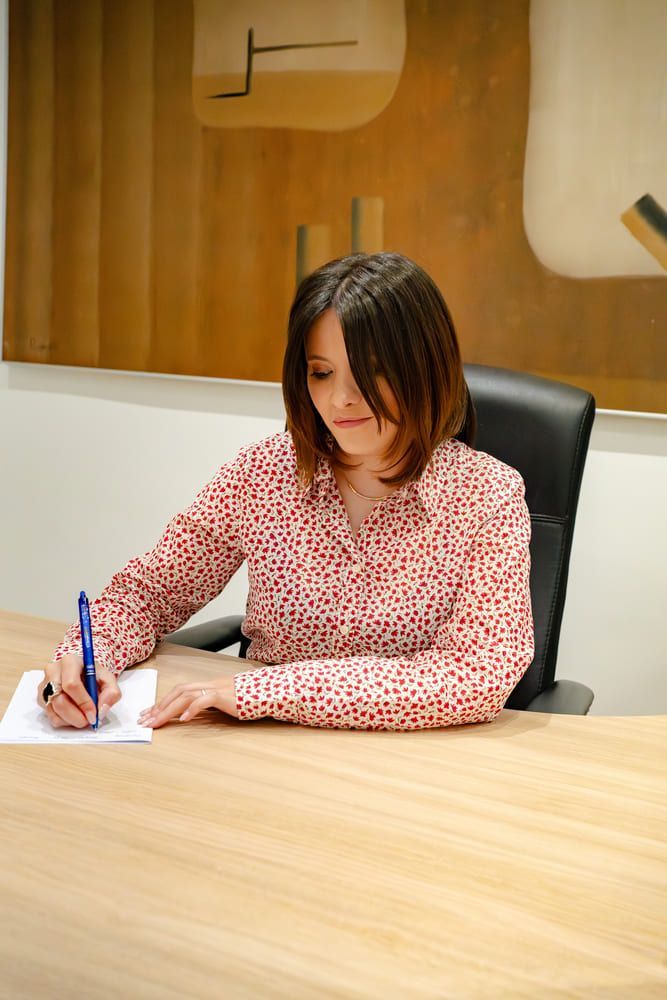 Mujer con camisa de lunares rojos, escribiendo en un escritorio, mirando hacia abajo. Silla de oficina negra, decoración de pared en tonos neutros.