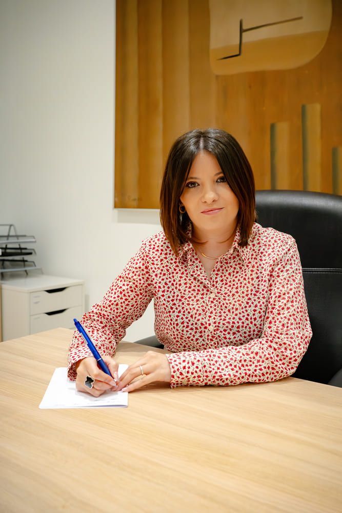 Mujer sentada a un escritorio, escribiendo en un papel. Camisa estampada roja y blanca, cabello castaño. Ambiente de oficina.