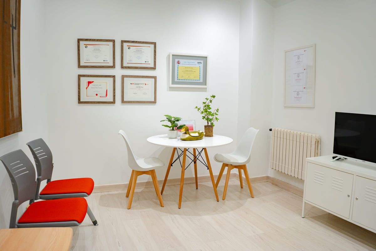 Sala de espera con paredes blancas, mesa, sillas, televisor y diplomas.