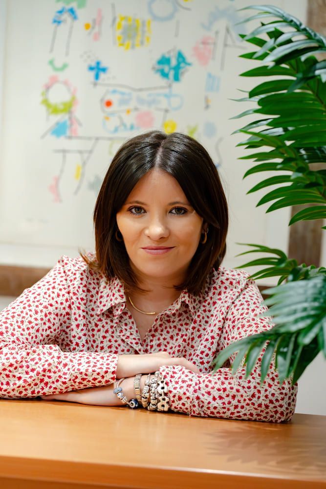 Mujer con camisa roja estampada, sentada a un escritorio de madera, mirando a la cámara.