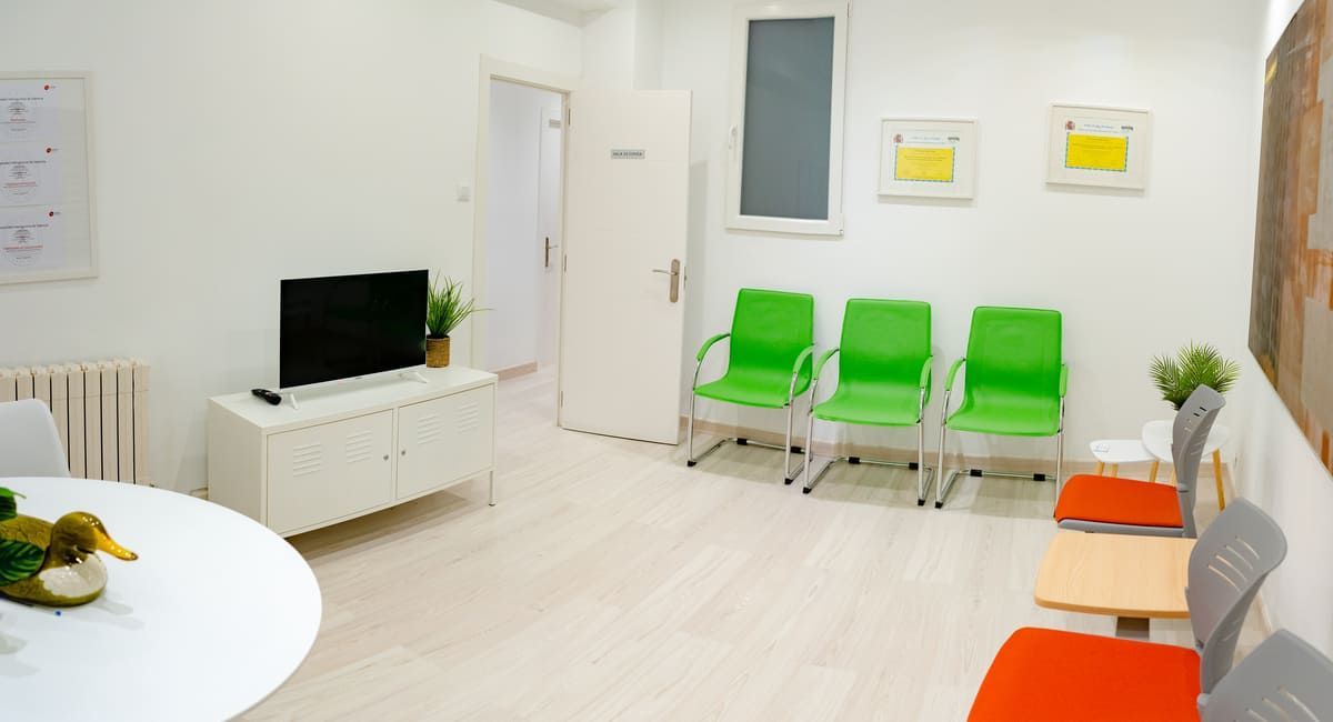 Sala de espera con paredes blancas, sillas verdes y naranjas, televisión y obras de arte.