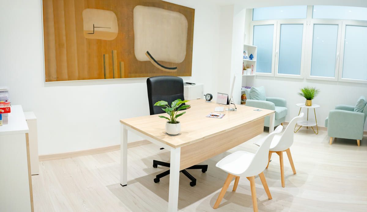 Una oficina bien iluminada con un escritorio, una silla y dos sillas blancas. En la pared cuelga una obra de arte abstracto.