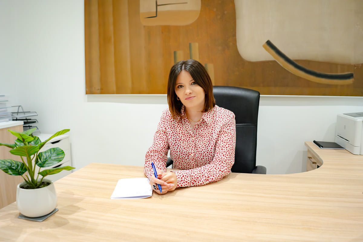 Una mujer con camisa estampada está sentada en un escritorio en una oficina, sonriendo, sosteniendo un bolígrafo.