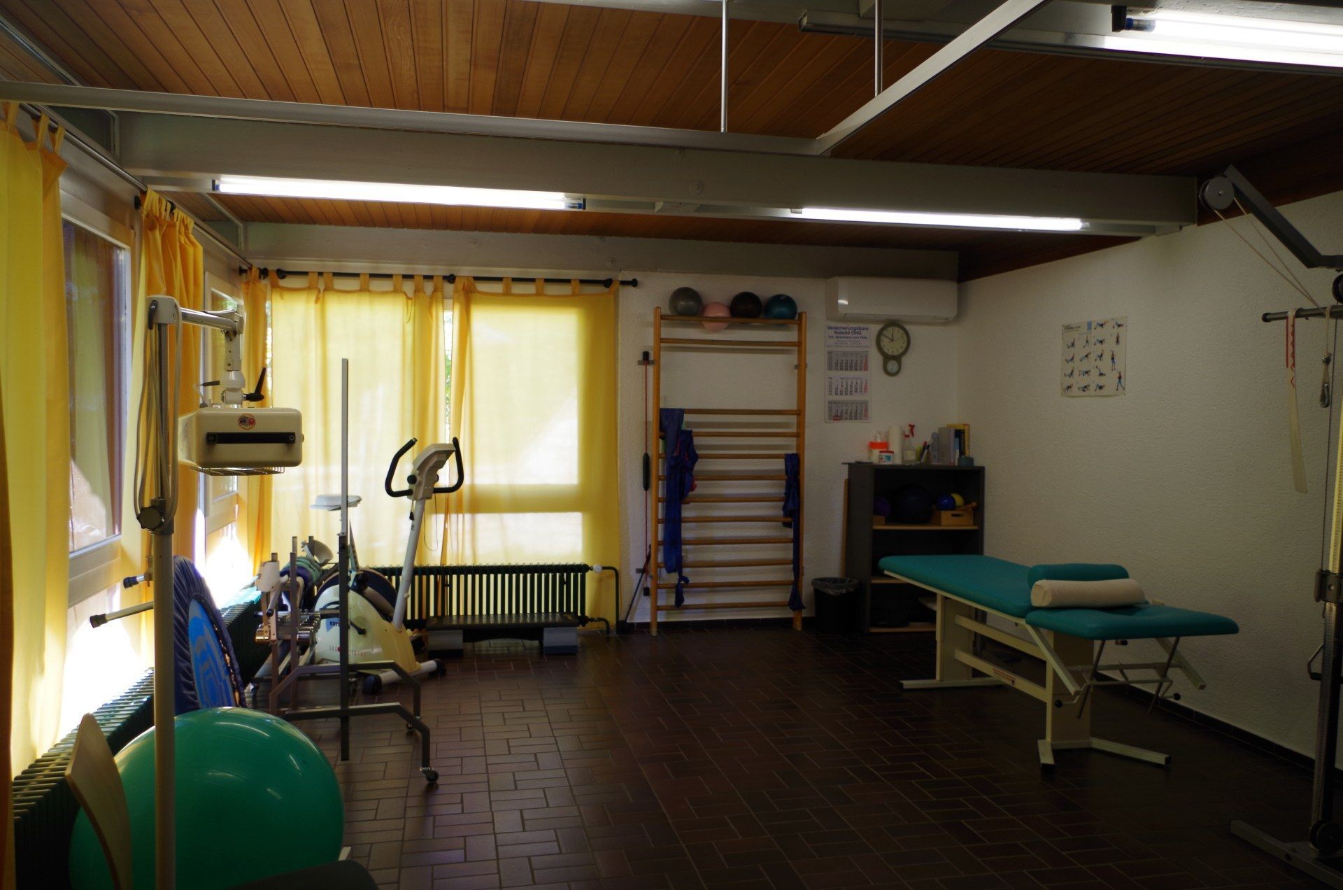 Rehabilitationsraum mit Trainingsgeräten und Behandlungstisch. Hellgelbe Vorhänge und Holzdecke.