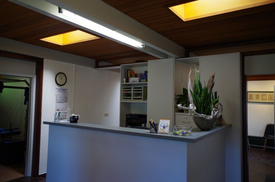 Empfangsbereich mit Schreibtisch, Pflanze und offenen Türen. Braune Decke, weiße Wände.