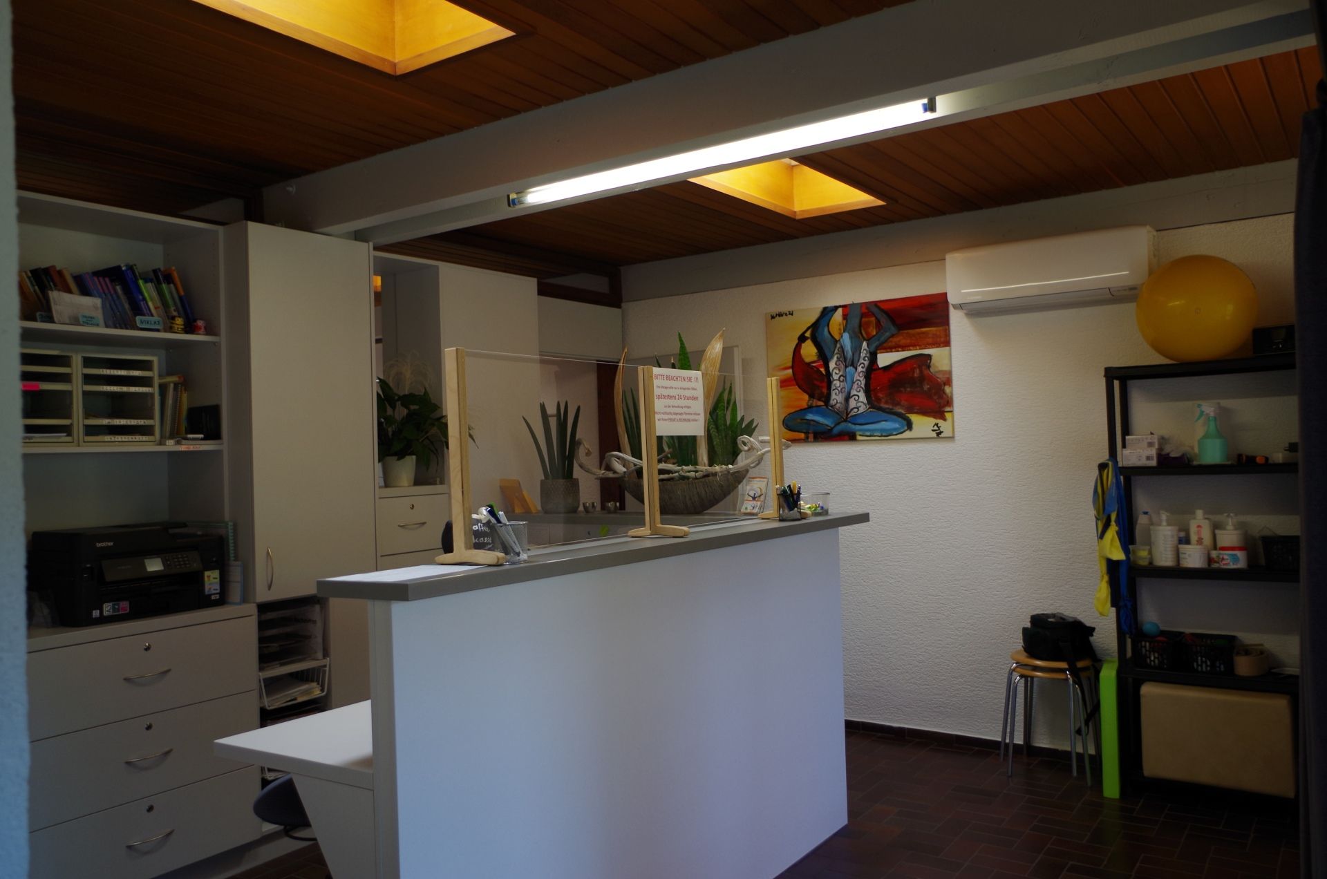 Empfangsbereich mit weißer Theke, Regalen und farbenfrohen Kunstwerken, gelber Gymnastikball.