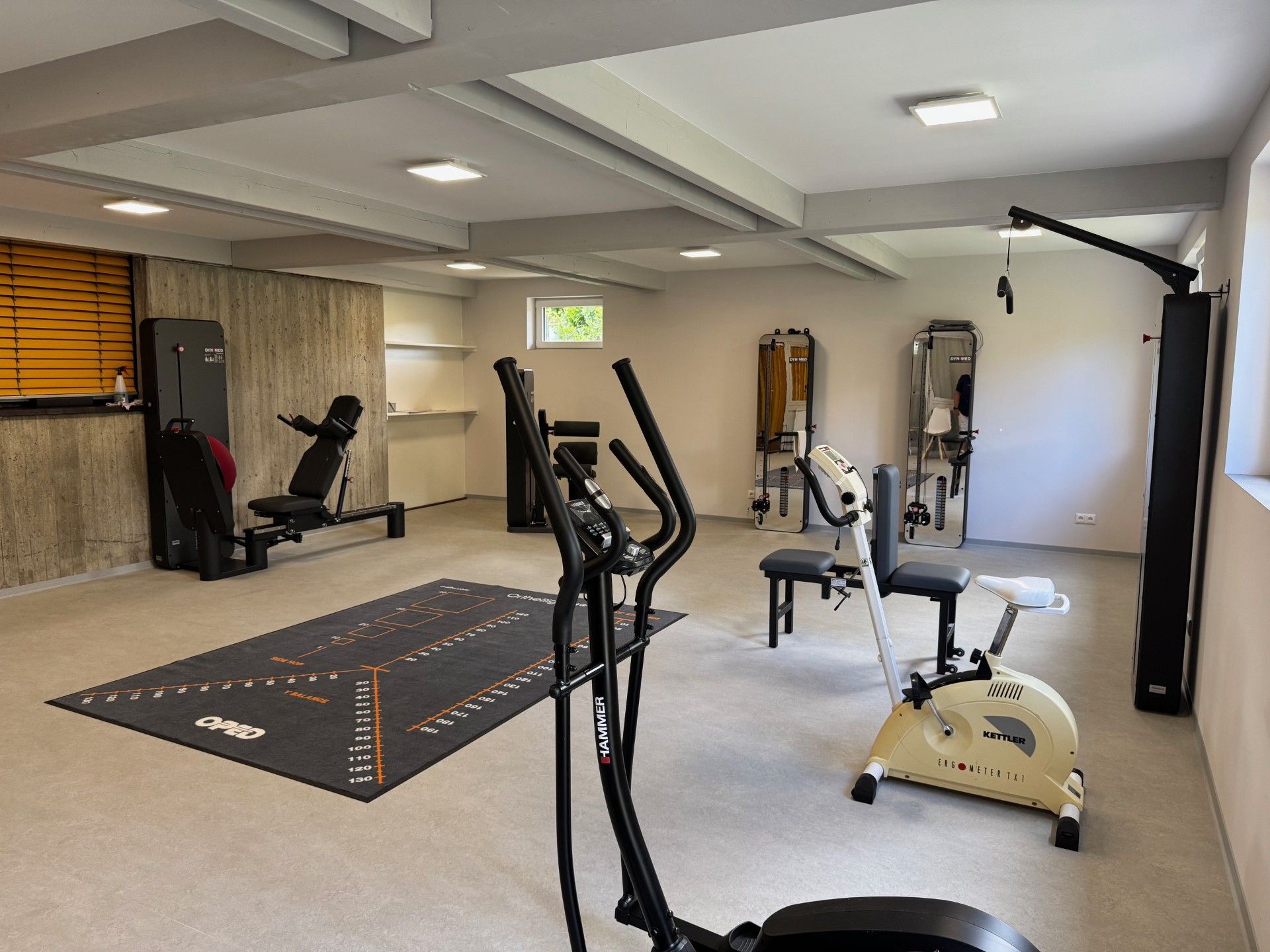 Heim-Fitnessstudio mit Trainingsgeräten auf hellem Boden. Zu den Geräten gehören ein Laufband, ein Heimtrainer und Kraftgeräte.