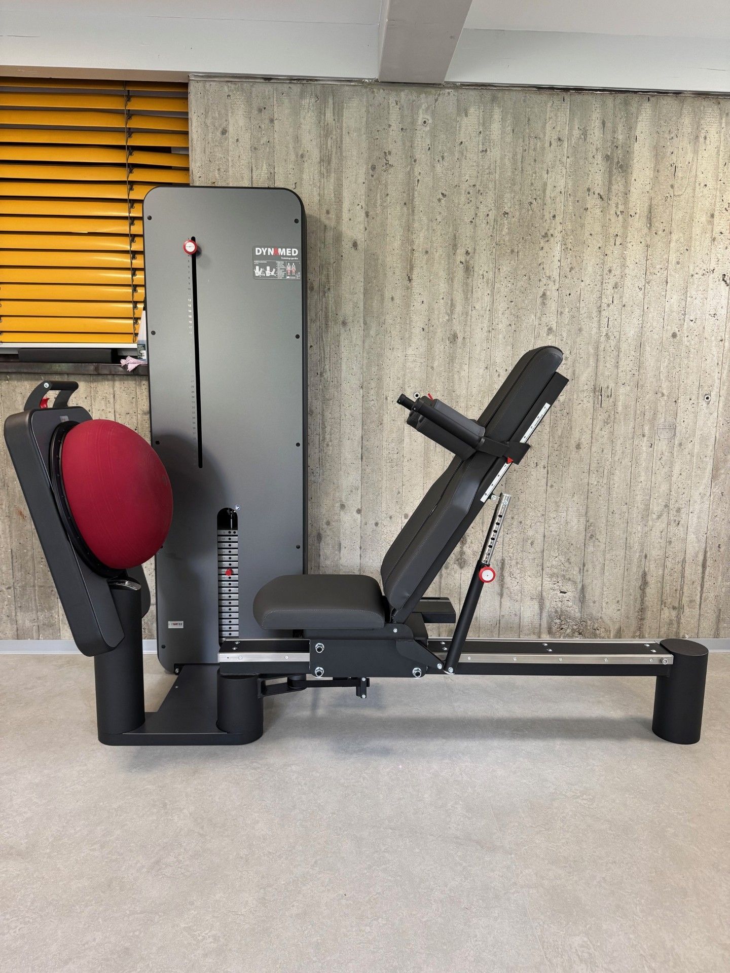 Eine graue Beinpresse mit einem roten Polster, vor einer Betonwand mit einer gelben Jalousie.