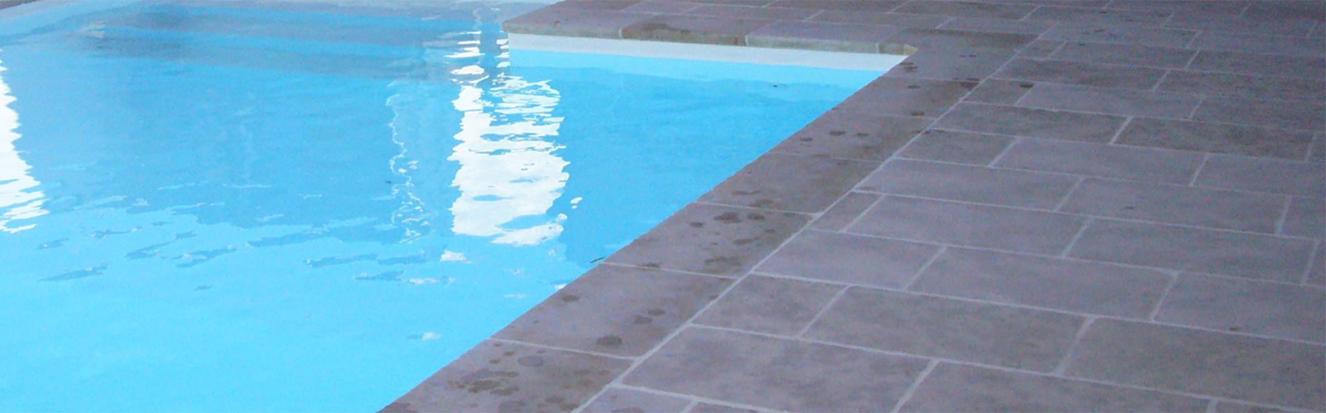 Piscine bleue jouxtant une terrasse carrelée grise où se reflètent les reflets de l'eau.