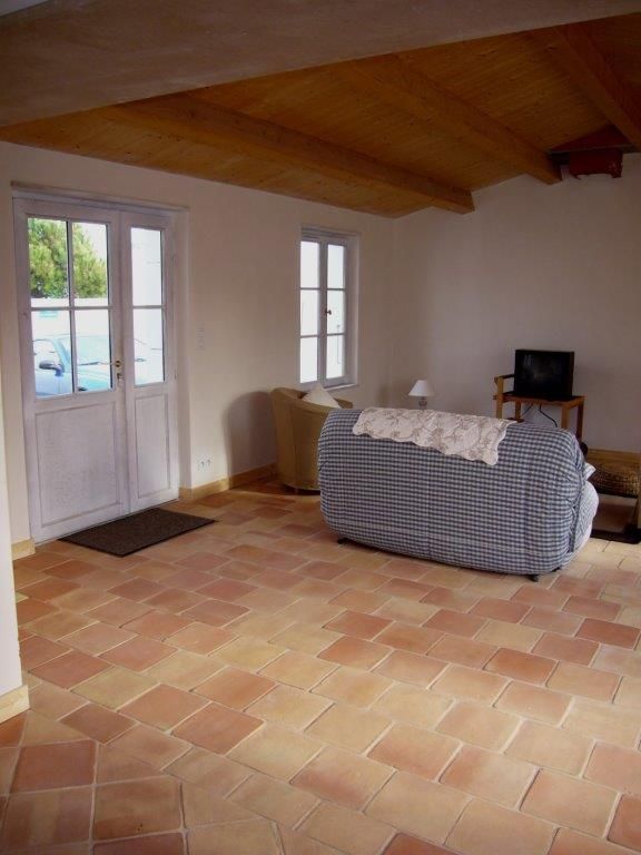 Salon vide avec sol en carrelage de terre cuite, plafond en bois, murs blancs et un canapé près de la porte.