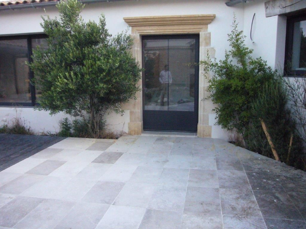 Entrée principale avec porte vitrée, encadrement en pierre et patio carrelé clair, flanqué de deux arbustes.