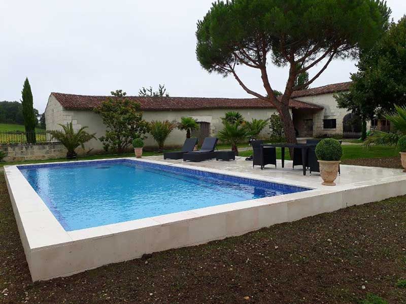 Piscine dans le jardin d'une maison, avec chaises longues, plantes en pot et un arbre sous un ciel couvert.