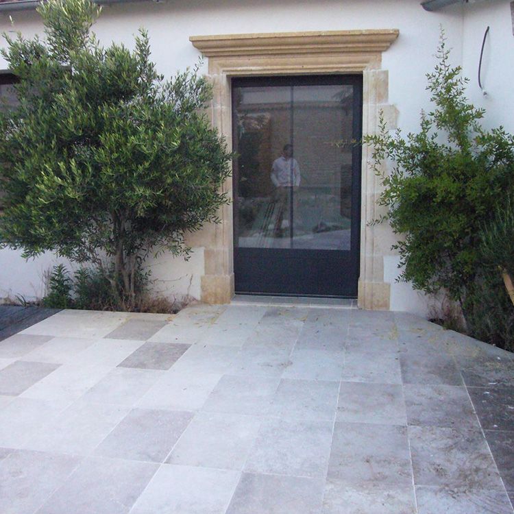 Une terrasse en pierre mène à une porte d'entrée vitrée encadrée de moulures beiges, flanquée d'arbustes verts.