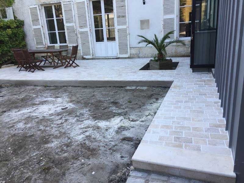 Patio avec allée en briques claires, jardin nu, table et chaises, et façade blanche de la maison avec portes-fenêtres