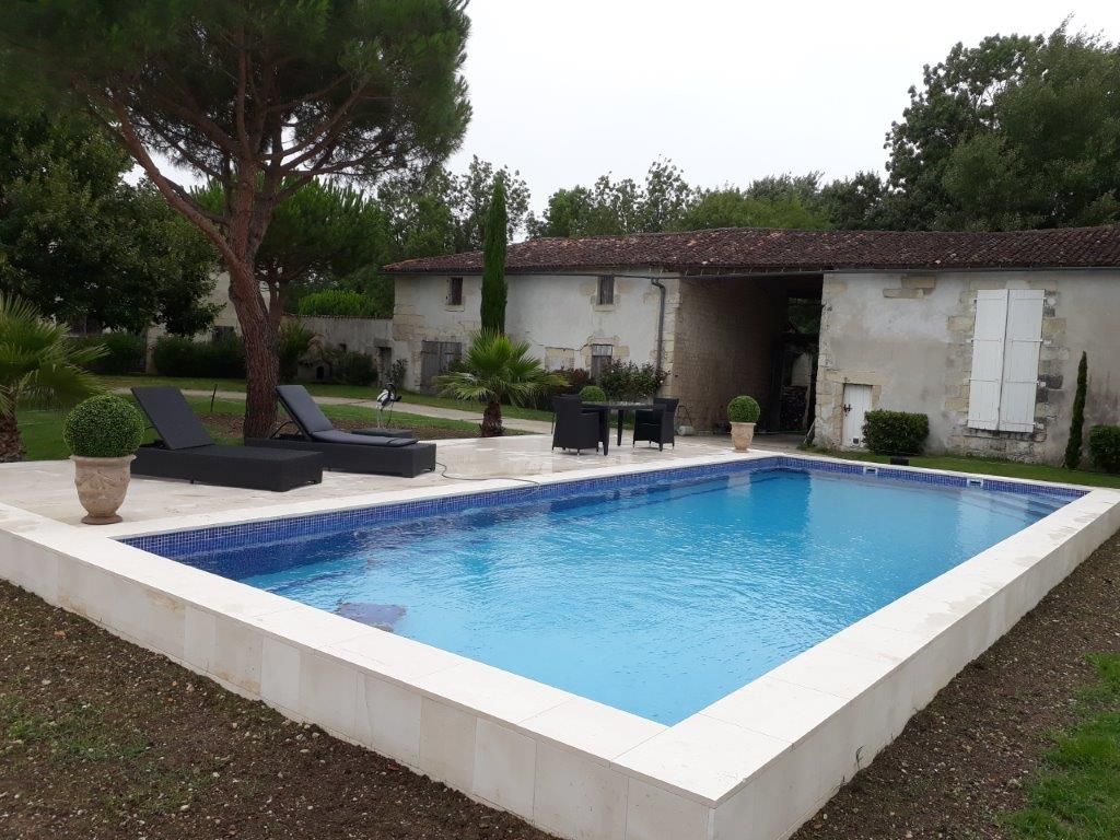 Jardin moderne avec piscine rectangulaire bleue, chaises longues, arbres et maison en pierre.
