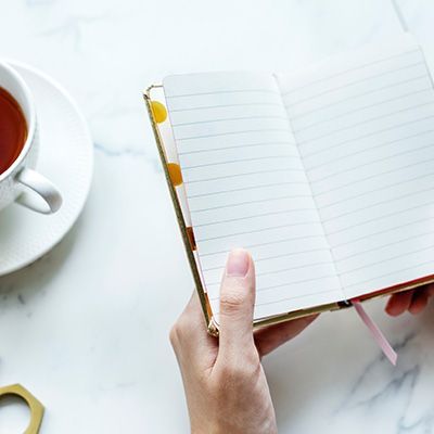 Main tenant un cahier à lignes ouvert à côté d'une tasse de thé sur un bureau en marbre blanc