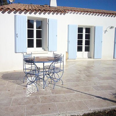 Terrasse avec table et chaises bleues devant une maison blanche aux volets bleu clair.