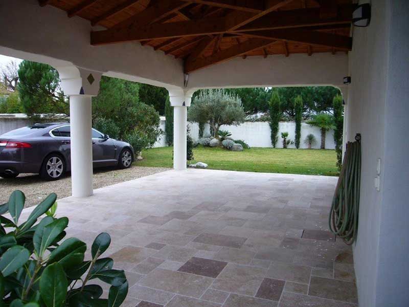 Terrasse carrelée couverte donnant sur une pelouse, avec une voiture garée et des colonnes blanches sous un toit en bois.