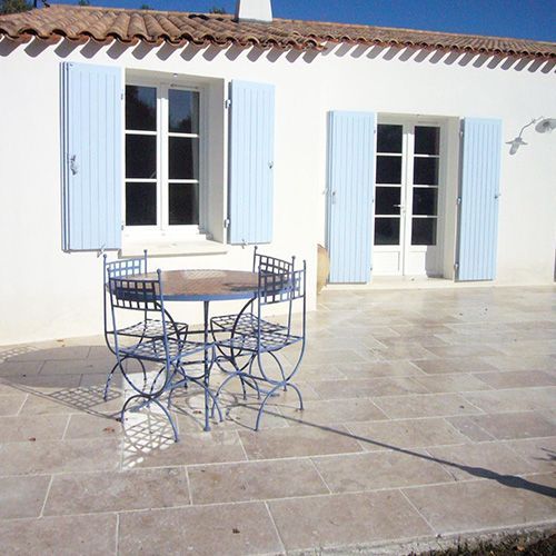Terrasse ensoleillée avec une table et des chaises en métal bleu devant une maison blanche aux volets bleus et aux portes-fenêtres