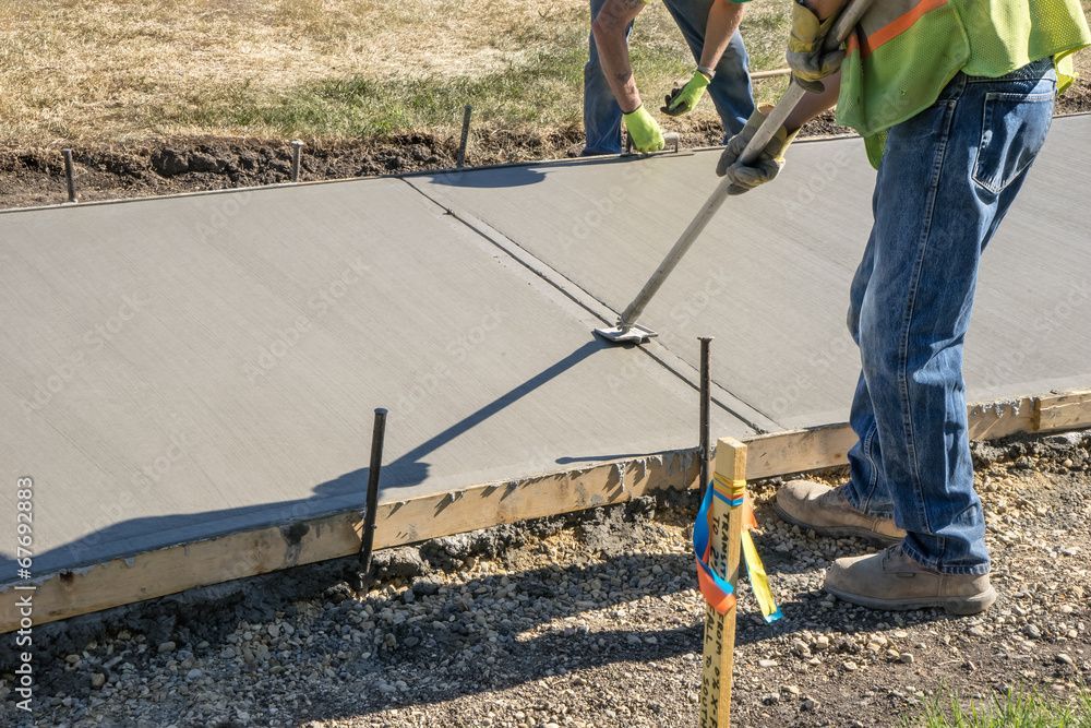 Arbeiter glätten mit einem langen Werkzeug den nassen Beton auf dem Gehweg neben einem frisch gegossenen Bordstein.