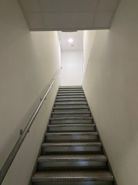 Escalier montant, marches et rampe en métal, murs blancs, plafond éclairé en haut.