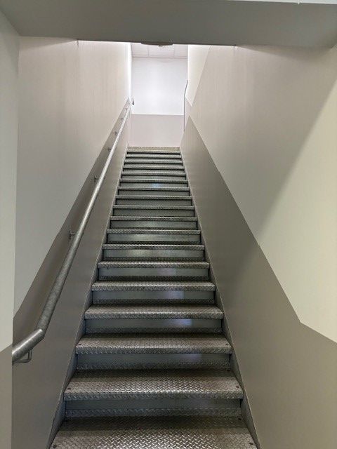 Un escalier métallique avec rampe monte entre des murs gris, menant à une ouverture lumineuse.