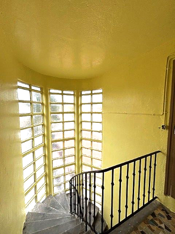 Intérieur jaune avec escalier en colimaçon, fenêtres en pavés de verre et balustrade noire.