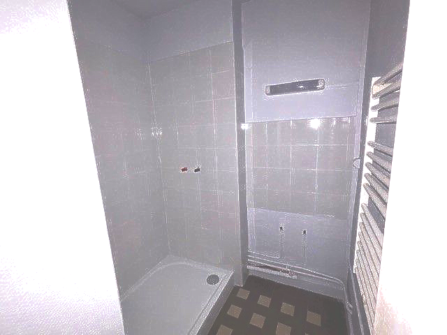 Petite salle de bain grise avec douche, chauffe-eau et murs carrelés.