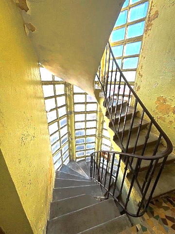 Escalier en colimaçon avec fenêtre, murs jaunes et rampe en métal. La lumière du soleil inonde l'espace par les fenêtres.