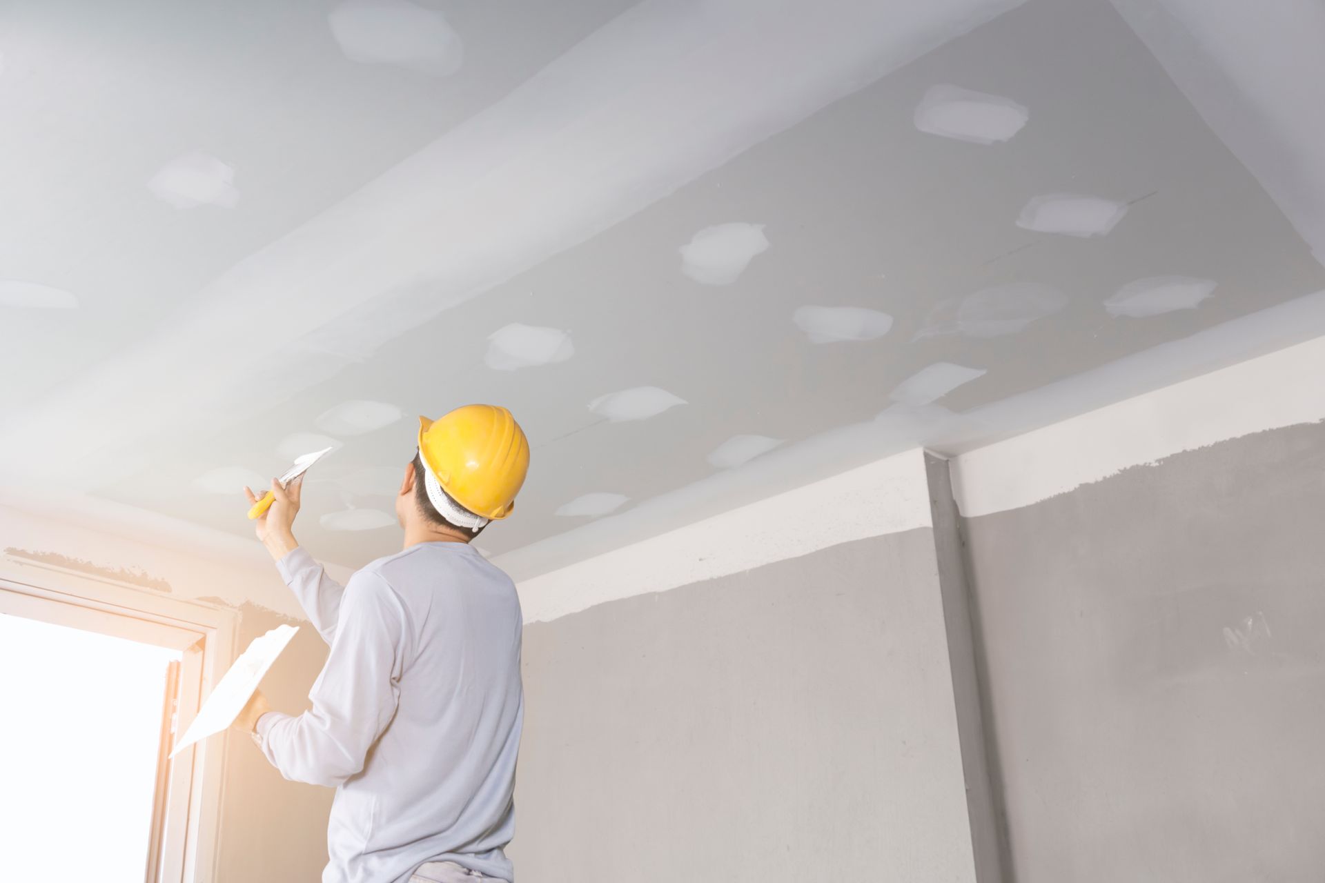 Un ouvrier portant un casque de chantier répare un plafond avec du mastic. La pièce est en plaques de plâtre.