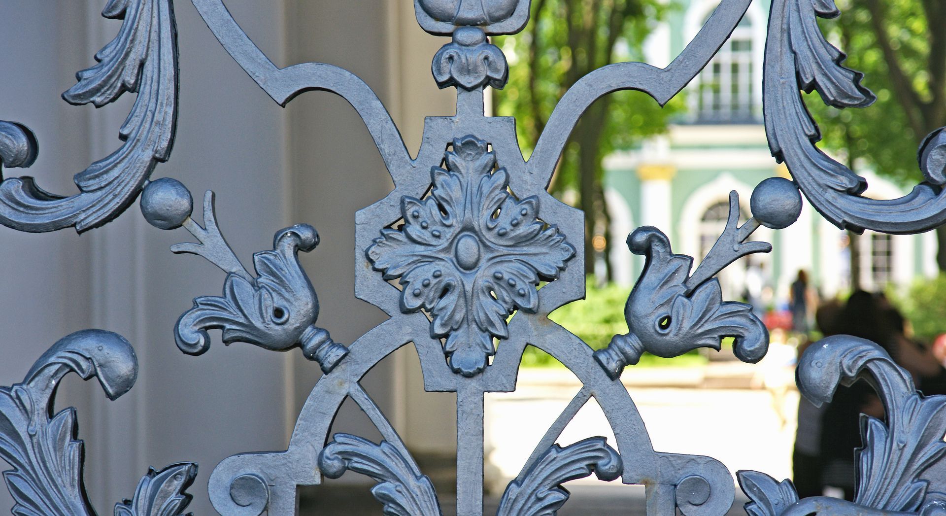 Clôture ornée en fer forgé argenté, avec un bâtiment et de la verdure visibles en arrière-plan.