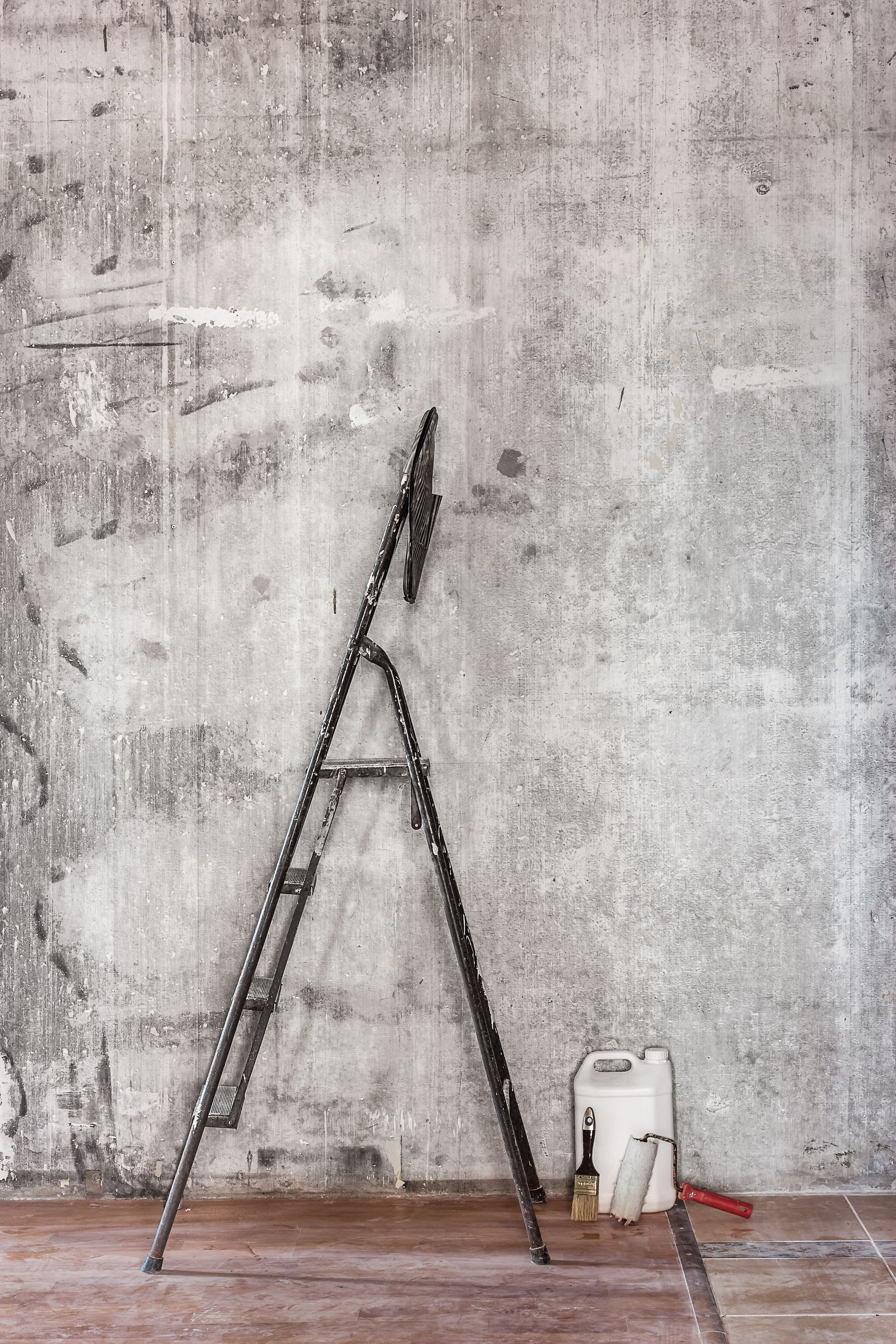 Une échelle appuyée contre un mur en béton usé, avec un récipient, une brosse et une scie au sol.