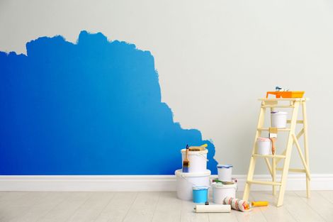 Mur partiellement peint en bleu, pots de peinture, rouleau, pinceaux et échelle sur un mur blanc.