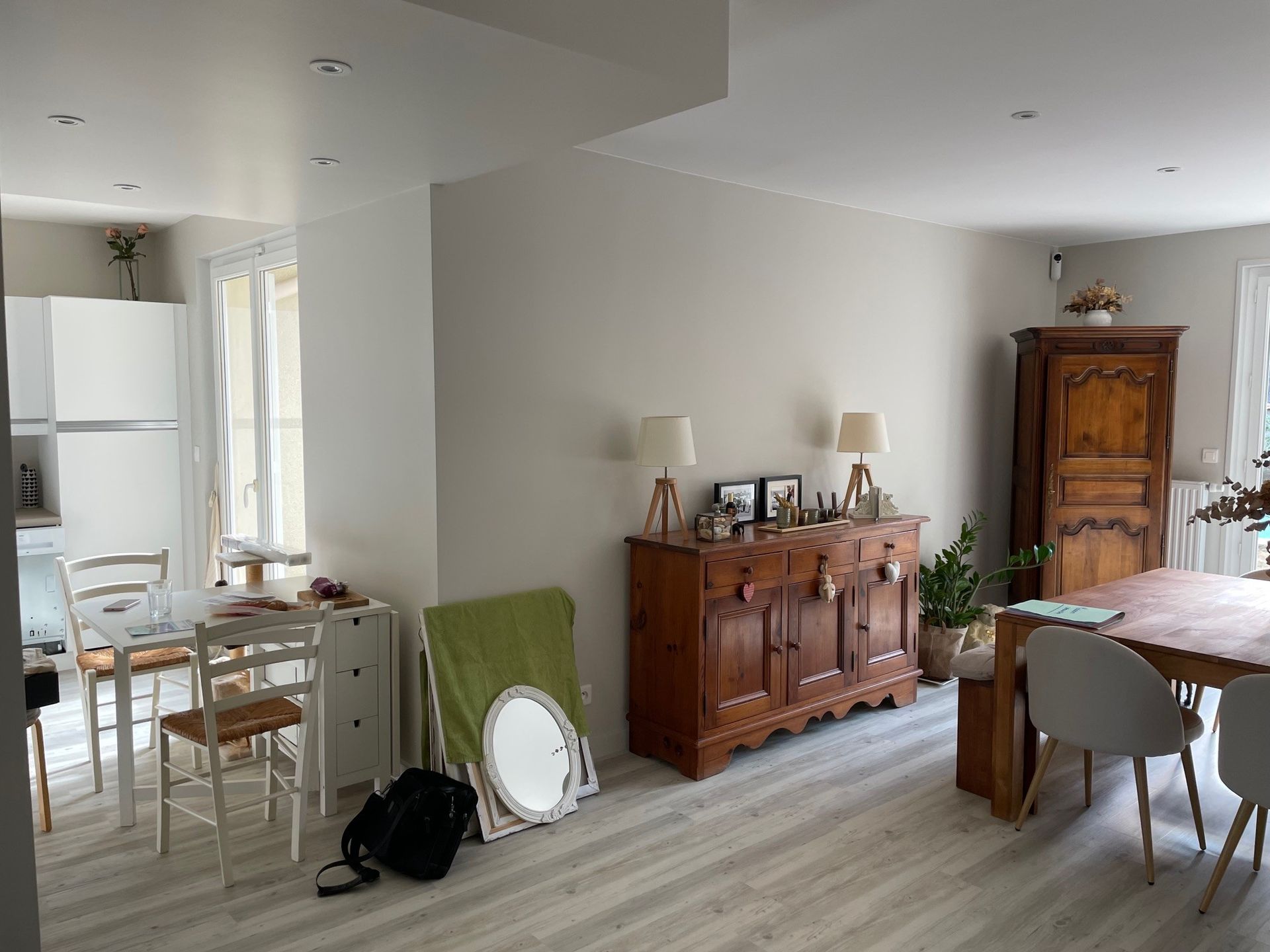 Intérieur meublé de meubles en bois, table à manger, chaises et cuisine visible. Murs gris et blancs, parquet.