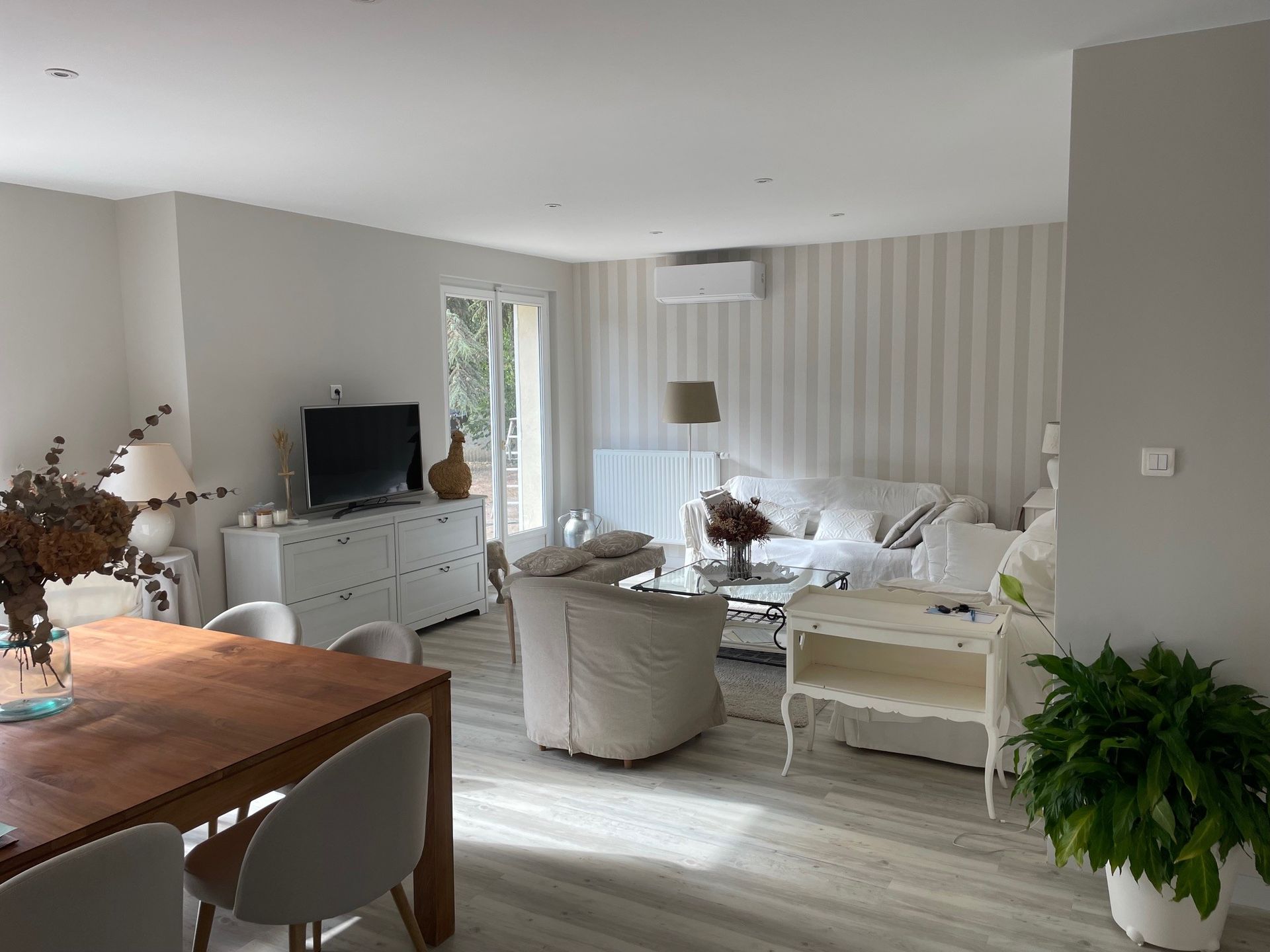 Salon avec murs clairs, papier peint à rayures, meubles blancs, table en bois et plantes d'intérieur.