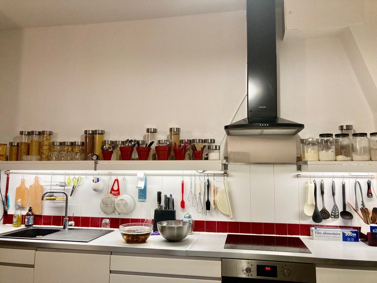 Cuisine avec placards blancs, touches de rouge, étagères ouvertes avec bocaux et ustensiles suspendus.