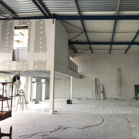 Intérieur d'un chantier de construction ; ouvrier sur une nacelle élévatrice, cloisons sèches, poutres apparentes, sol gris.