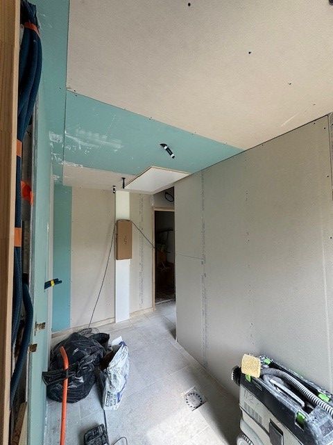 Couloir en travaux, plaques de plâtre installées aux murs et au plafond ; tons bleus et blancs ; outils et débris au sol.