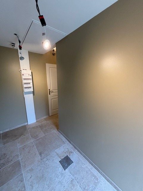 Couloir aux murs beiges, sol en pierre et porte blanche au fond. Éclairage sur rail au plafond.