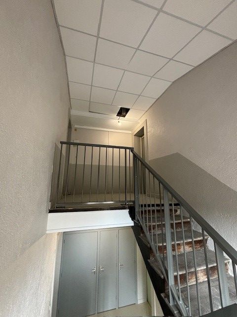 Cage d'escalier avec rampe et marches métalliques. Dalle de plafond endommagée laissant apparaître le luminaire. Murs blancs.