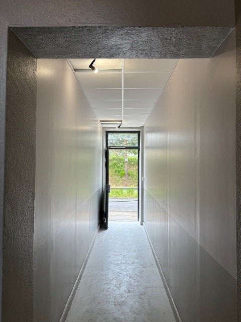 Couloir étroit aux murs et au plafond gris clair, menant à une porte ouverte donnant sur la verdure.