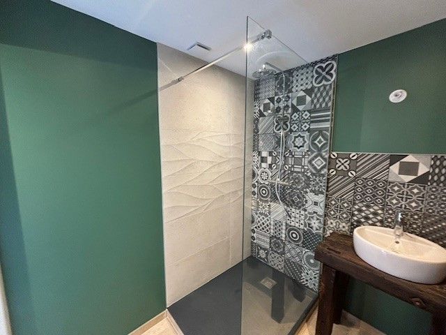 Salle de bains moderne avec douche en verre, murs verts, carrelage à motifs et meuble-lavabo en bois avec lavabo blanc.