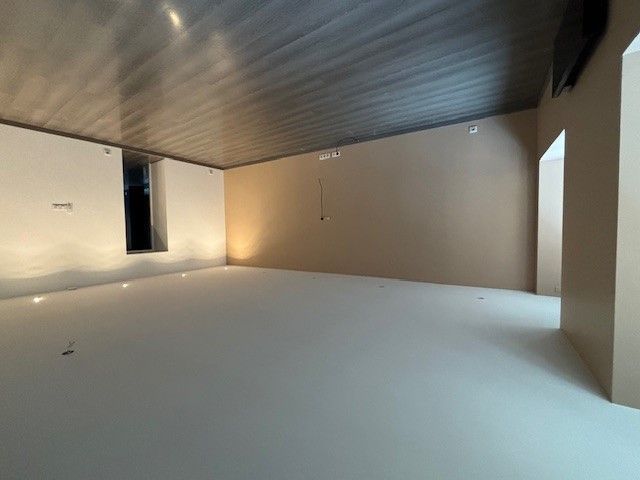 Pièce vide avec sol blanc, murs beiges et plafond sombre. Il y a une petite fenêtre sur la gauche.