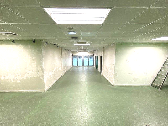 Long couloir vide, au sol vert et aux murs blancs. Des néons éclairent le plafond.